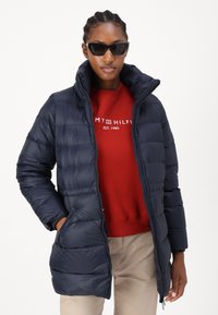 Navy puffer jacket with a high collar, zip closure, front pockets, and quilted design, worn over a red sweatshirt.