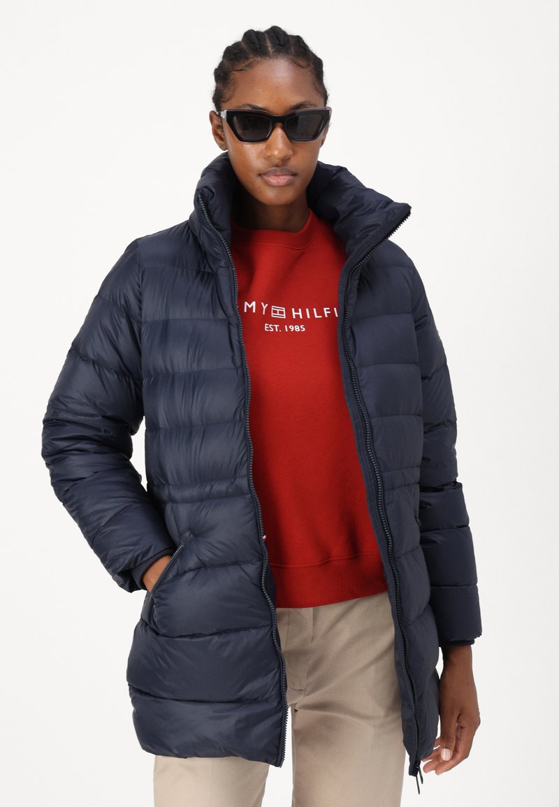 Navy Puffer-Jacke mit hohem Kragen, Reißverschluss, Fronttaschen und gestepptem Design, getragen über einem roten Sweatshirt.