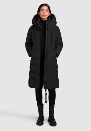 Vrouw in een lange zwarte gewatteerde jas met capuchon, zwarte leggings en zwarte enkellaarzen, staand tegen een effen lichte achtergrond.