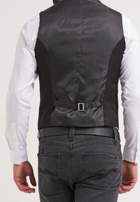 Schwarze Weste mit satinierter Oberfläche, mit einem einzelnen Schnallenriemen hinten, getragen über einem weißen Hemd und grauen Jeans.