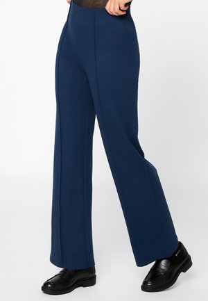 Persoon die marineblauwe, hoog getailleerde, wijde broek draagt en zwarte loafers, staand tegen een effen witte achtergrond.