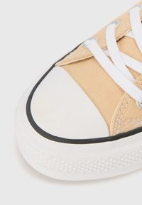 Beige lerret sneakers med hvit gummitå, svart kant og hvite snørebånd; mønstret såletekstur synlig langs kanten.