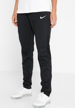 Personne portant un pantalon de sport noir Nike et des baskets blanches Nike, debout devant un fond blanc uni.