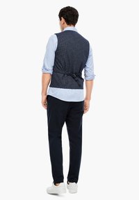 Marinefarbener, strukturierteter Weste mit einem taillierten Rücken, verstellbarem Riemen, getragen über einem hellblauen Hemd, kombiniert mit dunklen Hosen und weißen Sneakers.