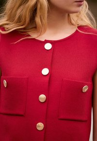 Top senza maniche lavorato a maglia rosso con tre bottoni dorati sul davanti e due tasche quadrate con dettagli rotondi dorati.