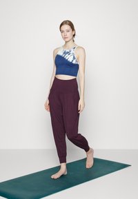 Sutiã desportivo em azul marinho com detalhes em tie-dye, combinado com calças harem soltas em roxo. Colocado sobre um tapete de yoga verde num fundo neutro.