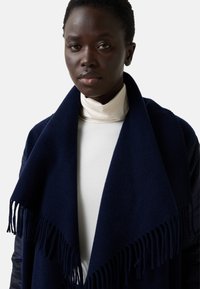 Námořnický vlněný šál s třásněmi, přehozený přes světle hrdlový top, s hladkou texturou a teplým vzhledem.