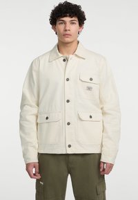 Cremfarbener Canvas-Jacke mit Knopfleiste, zwei Fronttaschen mit Klappen und einem kleinen Logo-Patch auf der Brusttasche.