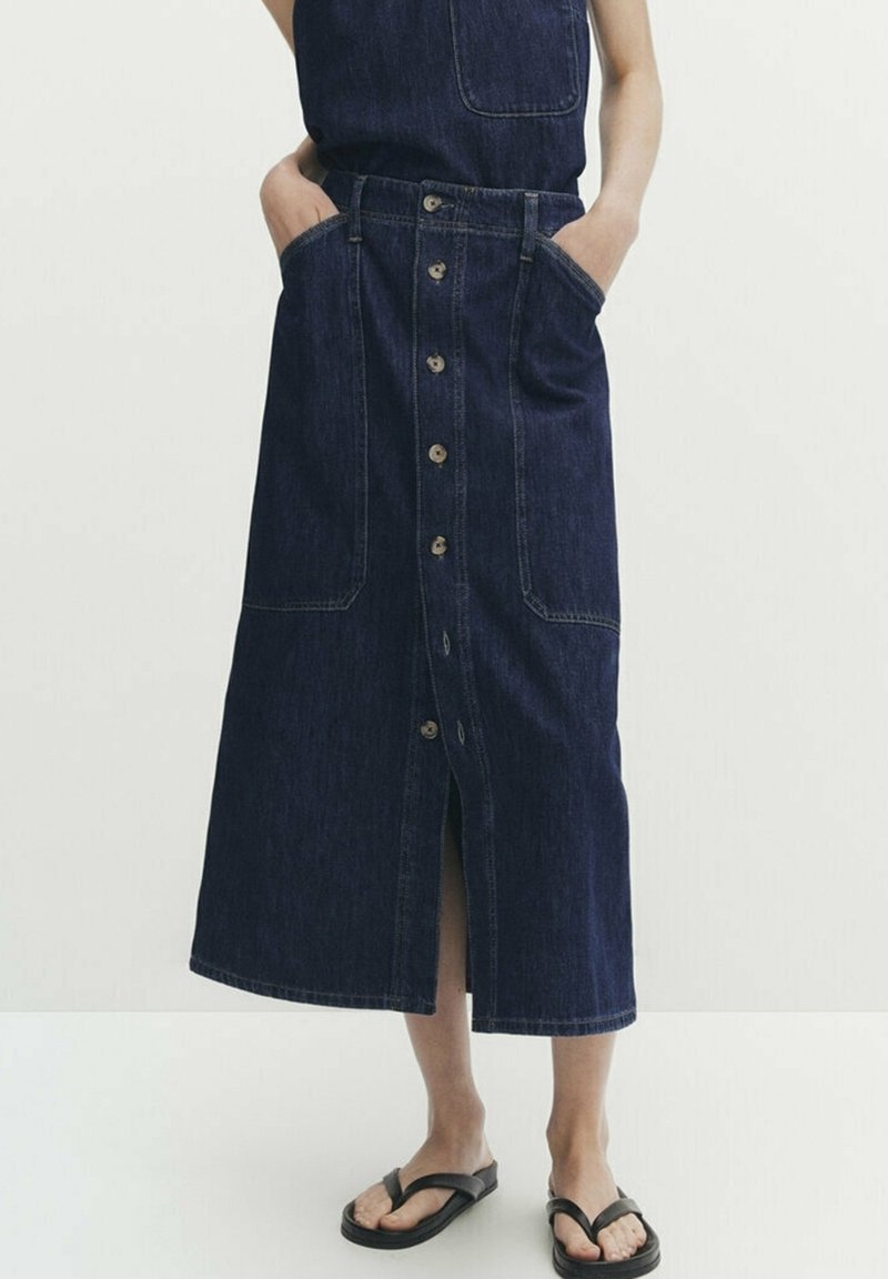 Femme portant une jupe en jean bleu foncé avec boutons sur le devant, poches profondes à l'avant et sandales noires, debout devant un fond uni.