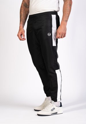 Zwarte sportbroek met witte zijstrepen, elastische tailleband en logo op de linker dij. Draagt witte sneakers met marineblauwe accenten.