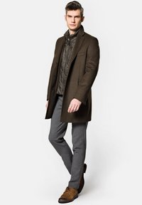 Brązowy wełniany płaszcz nałożony na szary pikowany wewnętrzny żakiet, w połączeniu z szarymi spodniami i brązowymi zamszowymi butami. Czysty design, dopasowany krój.