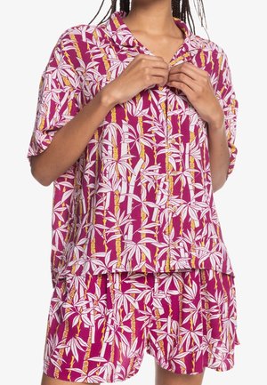 Osoba zapinająca koszulę w kolorze fioletowym z białym i żółtym wzorem liści bambusa, w zestawie z pasującymi szortami.