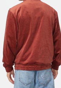 Rostfärgad, mjuk velourjacka med hög krage, elastiska muddar och fåll; bärs över ljusblå denimjeans med broderidetaljer.