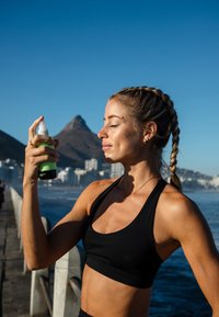 Femme aux cheveux tressés en tenue de sport noire aspergeant de la brume sur son visage près d'un front de mer avec des montagnes et des bâtiments en arrière-plan.