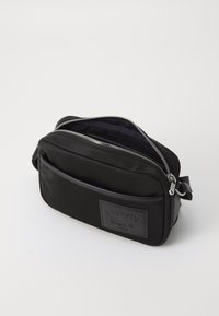 Borsa a tracolla nera in nylon con texture liscia, zip argentata e logo in rilievo sul fronte. Forma compatta e rettangolare con tracolla regolabile.