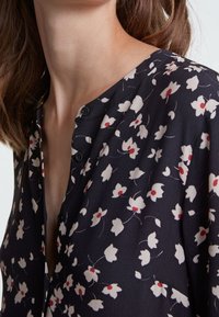 Kwiatowa bluzka w ciemnogranatowym kolorze z białymi i czerwonymi wzorami kwiatowymi. Ma dekolt w kształcie litery V z ozdobnymi guzikami oraz delikatną teksturę.