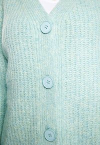 Cardigan tricotat de culoare bleu deschis, cu decolteu în V, trei butoane și o textură distinctivă. Butoanele sunt rotunde și au un luciu subtil.