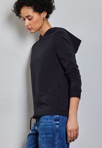 Schwarzer Hoodie mit Kordelzug, gerippten Bündchen und Saum. Kombiniert mit blauen Jeans. Einfaches Design vor einem neutralen Hintergrund.