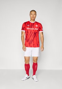 Maglia da calcio rossa con motivo astratto, dettagli bianchi, abbinata a pantaloni corti bianchi e calze rosse al ginocchio. Scarpe bianche con dettagli neri.