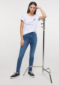 Balta kokvilnas T-kreklu ar zilu grafiku, pieguļošas zila džinsa bikses un tumši zilas sporta kurpes. Modelis stāv blakus hromētam fotostandam.