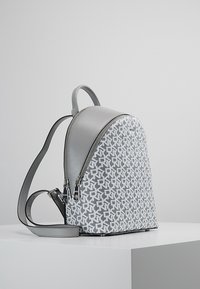 Grå faux läder ryggsäck med en rundad form, som har ett vitt geometriskt mönster, dubbla dragkedjor och justerbara remmar.