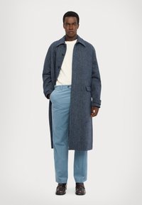 Donkerblauwe herringbone wollen jas met een notch kraag, knoopsluiting en twee zakken, gecombineerd met lichtblauwe broeken en zwarte schoenen.