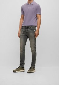 Polo viola con tre bottoni, jeans grigi in denim e sneakers verde oliva con dettagli neri. Il modello è in piedi davanti a uno sfondo semplice.