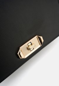 Schwarze Lederhandtasche mit einem strukturierten goldfarbenen Verschluss mit geometrischen Akzenten, umrandeter Naht und glatter Oberflächenbeschaffenheit.