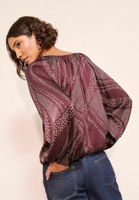 Bordeauxfarbene Bluse mit weißem gepunktetem Muster, lockeren Ärmeln und gerafftem Ausschnitt. Kombiniert mit Jeans aus Denim, die eine entspannte Passform zeigen.