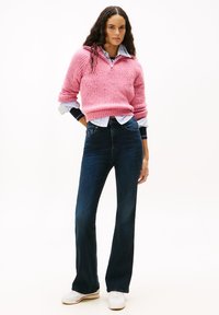Woman with long curly hair wearing a pink knit sweater over a striped shirt, dark blue jeans, and white sneakers, standing with hands in pockets.