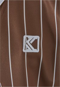 Brun vertikalt stribet stof har et hvidt broderet logo med et stiliseret bogstav "K" inden i en firkant. Materialet fremstår struktureret.