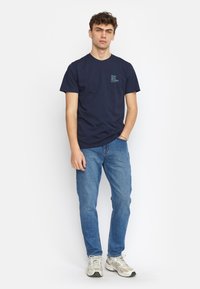 Navy blauwe katoenen t-shirt met korte mouwen, featuring een klein tekstontwerp op de linkerborst. Gecombineerd met lichtblauwe spijkerbroek en grijze sneakers.