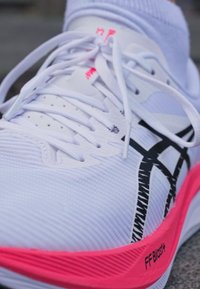 Närek av vit löparskor med svart logotyp, vita snörningar, rosa sula märkta "FF Blast+" och vit idrottssocka.