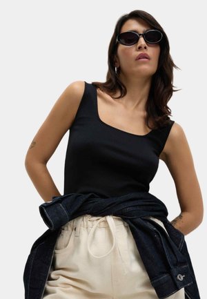 Débardeur noir ajusté avec un col carrément, pantalon beige taille haute, et une veste en denim foncé nouée autour de la taille.