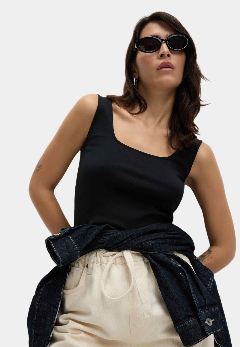 Schwarzes, enganliegendes Tanktop mit quadratischem Ausschnitt, beige, hochgeschnittene Hose und eine dunkle Jeansjacke um die Taille gebunden.