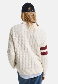 Off-white Strickpullover mit strukturierter Zopfmusterung, ausgestattet mit einem burgunderfarbenen Streifen am linken Ärmel und einem gerippten Saum. Lässiges Design.