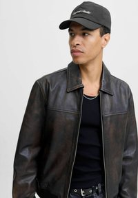 Brown leather jacket with a collared design, a zip front, and a textured finish; worn over a black ribbed tank top and accompanied by a black cap.