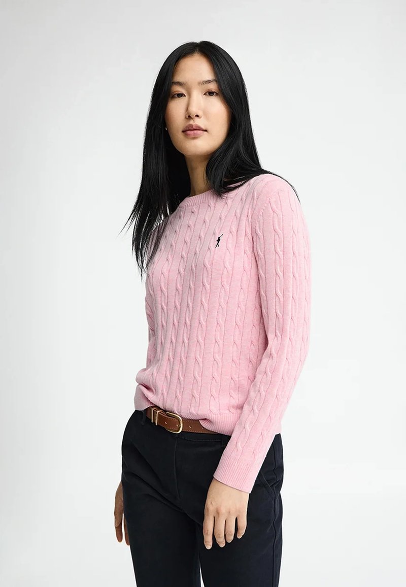 Joven con cabello largo y negro que lleva un suéter rosa de punto trenzado metido en pantalones negros con un cinturón marrón, de pie contra un fondo blanco.