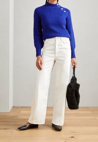 Pull bleu côtelé avec des accents de boutons, associé à un pantalon blanc à jambes larges et des ballerines noires. Le mannequin tient un sac à main noir texturé.