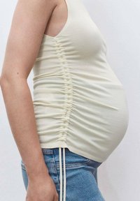 Top de maternidad beige claro sin mangas con fruncidos laterales, textura suave y diseño ajustado, combinado con jeans de mezclilla de lavado claro.