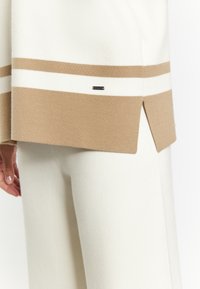 Cream sweater with beige stripes and side slits, paired with cream pants. Includes a small branded tag on the sweater. Soft knit texture.