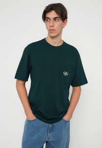 Temnozeleni bombažni T-shirt z okroglo odprtino, kratkimi rokavi in žepom na prsih, ki vsebuje večbarvno izvezeno logo.