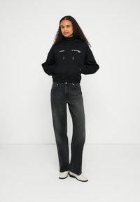 Sweatshirt preta com capuz e bolso frontal, jeans cinza de cintura alta e sapatos brancos. Modelo está em pé com as mãos nos bolsos, sobre um fundo simples.