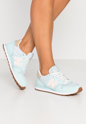 Nogi w jasnoniebieskich i białych butach New Balance z gumowymi podeszwami, stojące na białej powierzchni na tle jasnego, jednolitego tła.