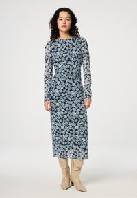 Maxi-jurk van zwarte transparante stof met een blauw bloemenpatroon. Lange mouwen, aansluitend lijfje en vloeiende rok. Gecombineerd met beige knielaarzen.