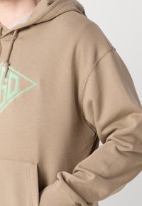 Felpa color beige con una texture morbida, caratterizzata da un logo verde menta a forma di diamante. Include una tasca frontale e un cappuccio con cordino.
