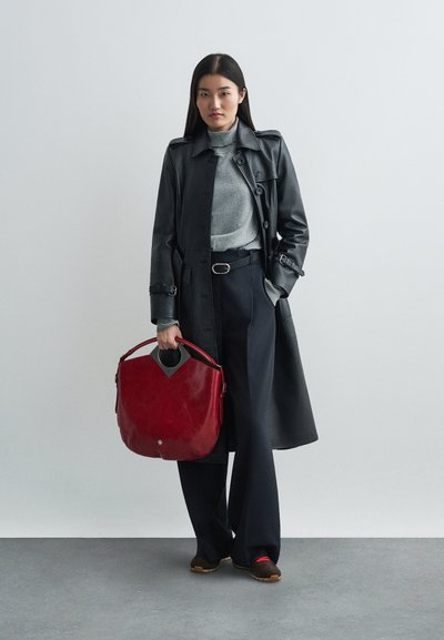 Manteau trench en cuir noir, col roulé gris, pantalons bleu marine et chaussures marron. Sac à main rouge avec un accent découpé unique, porté à la main.