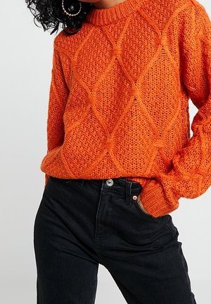 Femme portant un pull en maille torsadée orange avec des motifs en forme de losanges et un jean noir taille haute, main dans la poche.