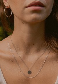 Silberne Creolen und geschichtete silberne Halsketten mit einem runden Anhänger, sichtbare Wassertropfen auf der Haut, sanfte natürliche Farbtöne.