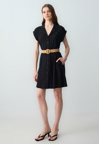 Abito camicia in lino nero con maniche corte, chiusura a bottoni sul davanti e tasche laterali, cinturato in vita con una cintura intrecciata color miele. Infradito nere.